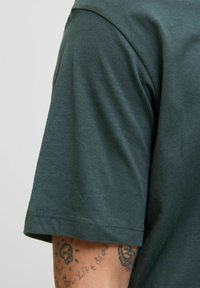 Mørkegrøn kortærmet T-shirt med en glat, blød tekstur. Har en rund halsudskæring og en syning i kanten, der delvist viser en tatoveret underarm.