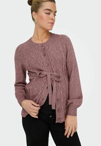 Adīta kardigāna mauve krāsā ar teksturētu dimanta rakstu, pogu aizdari, jostas saiti un ribotām piedurknēm. Modelis to valkā virs melnajām biksēm.