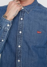Sininen farkkupaitakuvio, jossa on terävä kaulus, valkoiset napit ja pieni punainen Levi's-logo rinnassa.