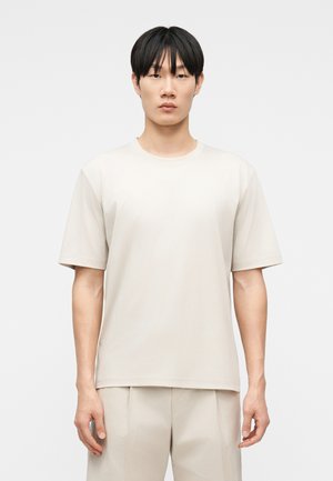 Jeune homme debout face à l'avant, portant un T-shirt à manches courtes uni beige clair et un pantalon beige clair assorti.