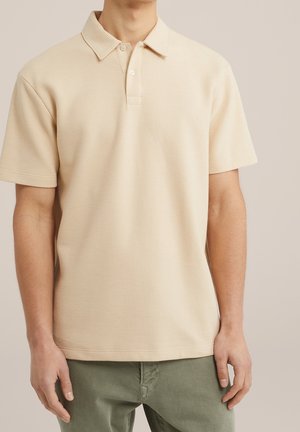 Beiges kurzärmeliges Poloshirt aus strukturiertem Baumwollstoff mit einem Kragen und einer dreiknöpfigen Blende, kombiniert mit grüner Hose.