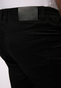 Svarta byxor med resårmidja, utrustade med en grå läderlapp märkt "BOSTON PARK" och en bakficka med synliga sömnadsdetaljer.