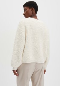 Vrouw met kort haar in een gestructureerde witte gebreide trui en lichtbeige broek, zijwaarts kijkend tegen een effen achtergrond.