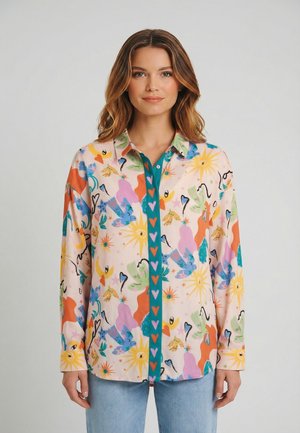 Donna che indossa una camicia colorata con bottoni, decorata con motivi astratti di sole, cuori e animali, abbinata a jeans azzurri, in piedi davanti a uno sfondo semplice.