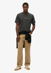 Camiseta gris de algodón con logo, pantalones beige y zapatillas marrón claro. Suéter de cuadros verde oscuro atado a la cintura.