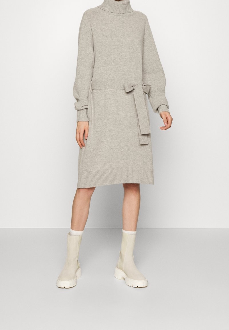 Femme portant une robe à col roulé tricotée gris clair avec ceinture nouée et bottes épaisses beige mi-mollet sur fond uni.