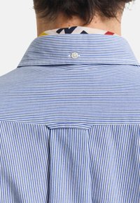 Camisa abotonada con finas rayas azules y blancas, que presenta un pequeño pliegue en la parte posterior y un colorido cuello con estampado visible en el cuello.