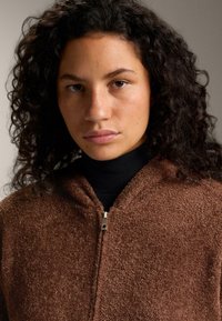 Hnedá bunda na zips vyrobená z textúrovaného materiálu, s vysokým golierom, lesklým zipsom a jemným, chlpatým vzhľadom.