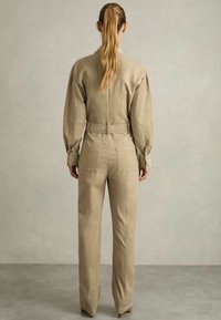 Salopetă bej, cu mânecă lungă, cu talie accentuată printr-un brâu, cu o croială ajustată și buzunare laterale. Materialul are o textură netedă și un design structurat.