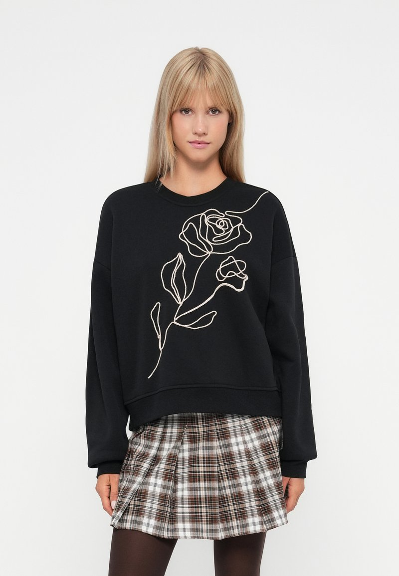 Joven mujer vestida con un sudadera negra que tiene un dibujo lineal blanco de una rosa y una falda de cuadros marrones con pliegues, de pie frente a un fondo blanco.