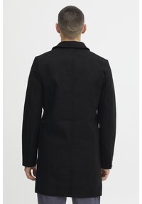 Schwarzer Wollmischfeldmantel mit taillierter Silhouette, Reverskragen und dreiknöpfigen Manschetten, mit glatter Textur und klaren Linien.