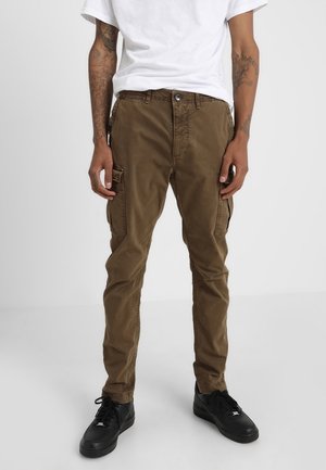 Mand iført brune cargobukser, sorte sneakers og en hvid t-shirt, stående med arme afslappet langs siderne på en ensartet lys baggrund.