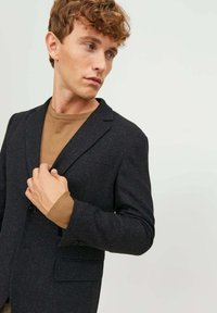 Blazer scuro e strutturato con un solo bottone, rever a punta e tasca laterale. Indossato sopra un maglione marrone, che mette in mostra colori sovrapposti.