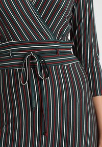 Šaty z pruhované látky v zelené, bílé a červené barvě; mají překládaný design s uvázaným páskem v pase a rukávy o délce tři čtvrtě.