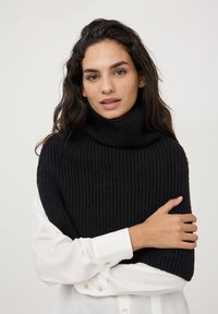 Poncho nero lavorato a maglia con colletto alto, caratterizzato da una texture a costine. Indossato sopra una camicia bianca con dettagli a bottoni sui polsini.