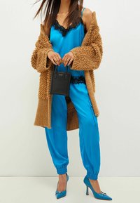 Frau trägt ein blaues Satin-Camisole und eine Hose mit schwarzem Spitzenbesatz, eine beigefarbene grobstrickige Strickjacke, blaue Absätze und hält eine kleine schwarze Handtasche.