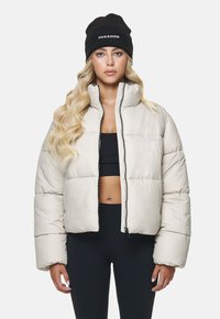 Puffige, kurze Jacke in Off-White, mit hohem Kragen und Reißverschluss. Kombiniert mit einer schwarzen Mütze sowie passendem schwarzen Oberteil und Hose.