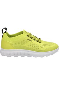 Zapatilla deportiva amarilla con parte superior de malla, textura circular, acentos negros y una suela blanca con un patrón de olas para amortiguación.