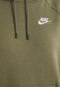Oliwkowa bluza z kapturem wykonana z miękkiego materiału, z przednią kieszenią i czarnymi troczkami, z białym logo Nike wyhaftowanym na piersi.