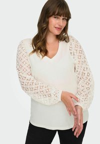 Hvit topp med V-hals og teksturerte blondermer i et blomsterdekor, kombinert med svarte bukser. Stoffet ser mykt og lett ut.