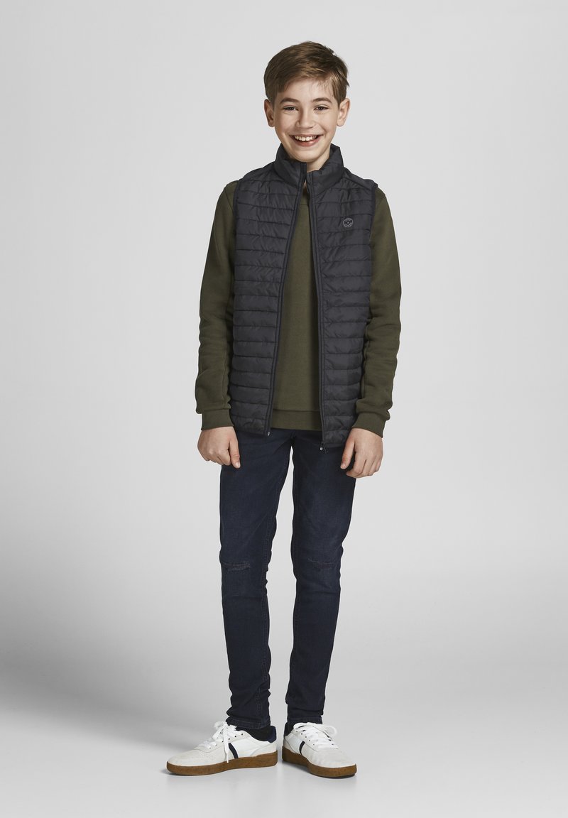 Zwarte gewatteerde vest met hoge kraag, olijfgroen shirt met lange mouwen, donkerblauwe jeans en witte sneakers met tan accenten, staande pose.