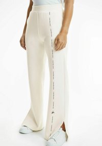 Off-white vida byxor med sidoflikar och upprepade "Calvin Klein Jeans"-logotyper längs ytterseamna, bärs med vita sneakers.