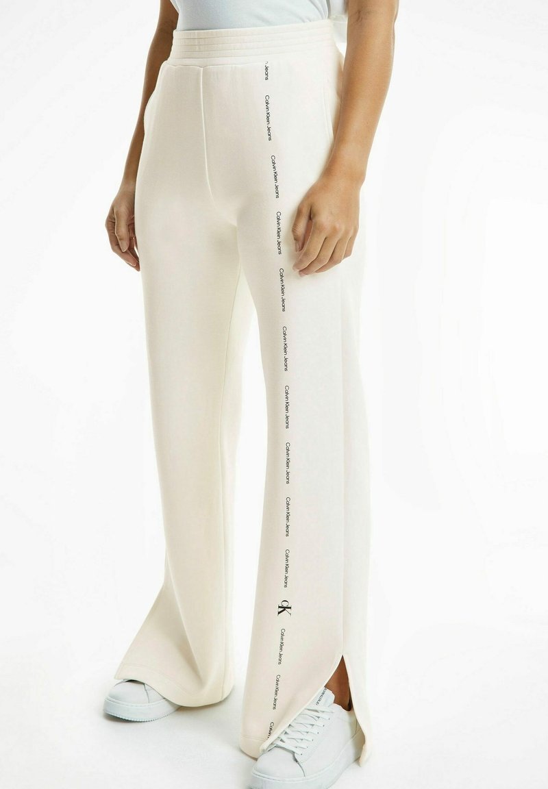 Off-white vida byxor med sidoflikar och upprepade "Calvin Klein Jeans"-logotyper längs ytterseamna, bärs med vita sneakers.