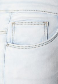 Tejido de mezclilla de color azul claro con un bolsillo trasero. Presenta costuras doradas a lo largo de las costuras y un detalle de botón sobre el bolsillo.