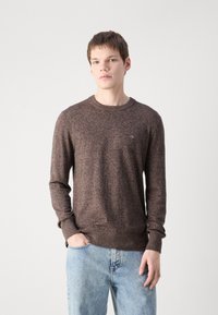 Brązowy sweter dzianinowy z okrągłym dekoltem, długimi rękawami i ściągaczami przy mankietach. Subtelna tekstura z małym logo na piersi, w połączeniu z jasnoniebieskimi dżinsami.