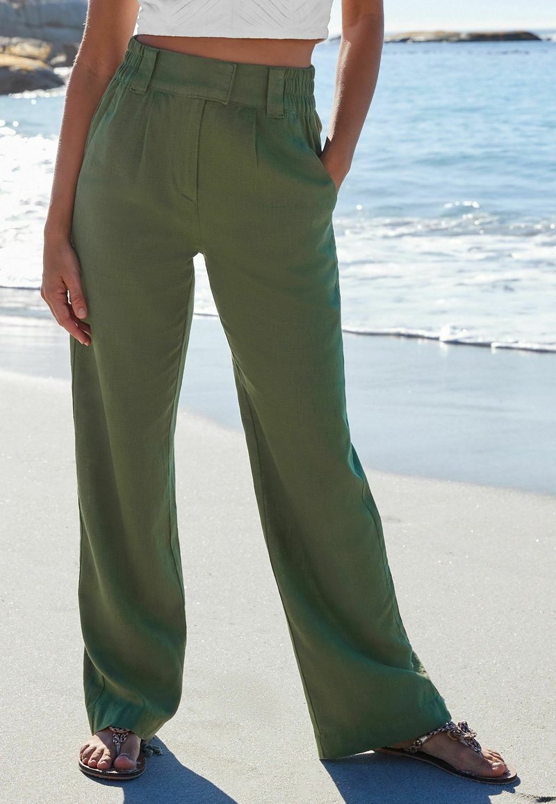 Pantalons larges verts en tissu léger, avec un devant plissé et une taille élastique, associés à des sandales plates sur une plage de sable.
