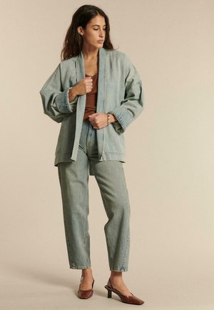 Kobieta ubrana w za dużą jasnoniebieską kurtkę jeansową i spodnie, brązowy top oraz brązowe sandały na obcasie z paskiem na pięcie, stojąca na beżowym tle.