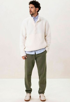 Pull en polaire blanc cassé avec un col zippé au quart, porté sur une chemise bleu clair, associé à un pantalon vert olive et des baskets blanches.