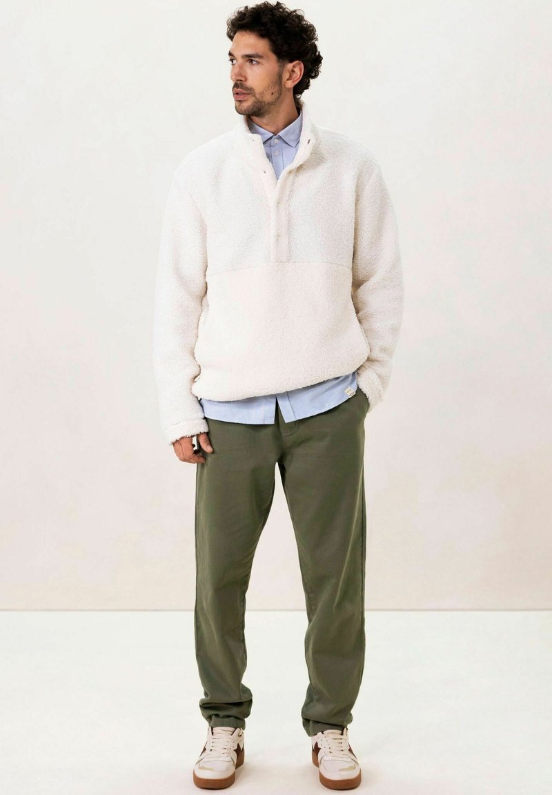 Pull en polaire blanc cassé avec un col zippé au quart, porté sur une chemise bleu clair, associé à un pantalon vert olive et des baskets blanches.