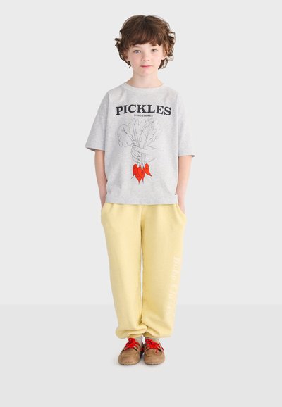 Enfant aux cheveux bouclés portant un t-shirt gris "PICKLES", un pantalon de survêtement jaune et des chaussures marron avec des lacets rouges, debout les mains dans les poches.