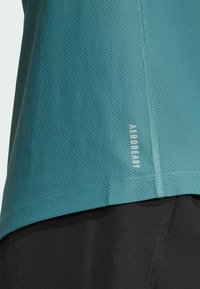 Tirkizna, teksturirana sportska majica od prozračnog materijala, s logotipom i detaljima šivanja. Uparena s crnim šortsevima.