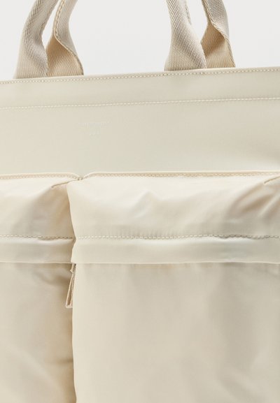 Μπεζ τοτ τσάντα με δύο επενδυμένες μπροστινές τσέπες και διπλές υφασμάτινες λαβές, με διακριτική ραφή και μικρό ανάγλυφο λογότυπο κοντά στην πάνω άκρη.