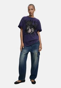 Camiseta morada de gran tamaño con un diseño gráfico negro texturizado, combinada con vaqueros azules de corte holgado, con bolsillos de parche y un acabado liso.