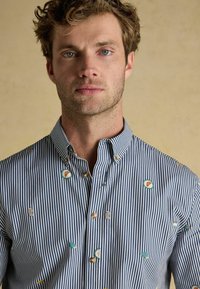 Košeľa s gombíkmi, ktorá sa vyznačuje modro-bielymi prúžkami a vyšívanými detailmi v rôznych tvaroch a farbách. Textúrovaná bavlnená tkanina.