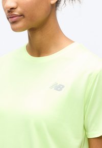 Κοντινό πλάνο ενός ατόμου που φοράει αθλητική μπλούζα New Balance ανοιχτού πράσινου χρώματος, με το λογότυπο στο αριστερό μέρος του στήθους και μέρος του προσώπου και του λαιμού να είναι ορατά.