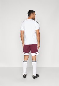 Heren sportoutfit bestaande uit een witte korte mouwen shirt, bordeauxrode shorts met witte strepen, witte kniehoge sokken en zwarte voetbalschoenen.