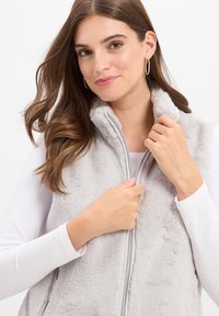 Szary kamizelka z futra syntetycznego z wysokim kołnierzem i zamek błyskawiczny z przodu. Modelka ma na sobie białą bluzkę z długim rękawem, co eksponuje teksturę i kolor kamizelki.