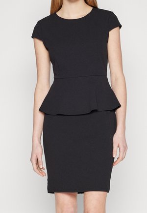 Kvinde iført en tætsiddende sort kjole med korte ærmer og peplum-detalje ved taljen, stående mod en ensfarvet lys baggrund.
