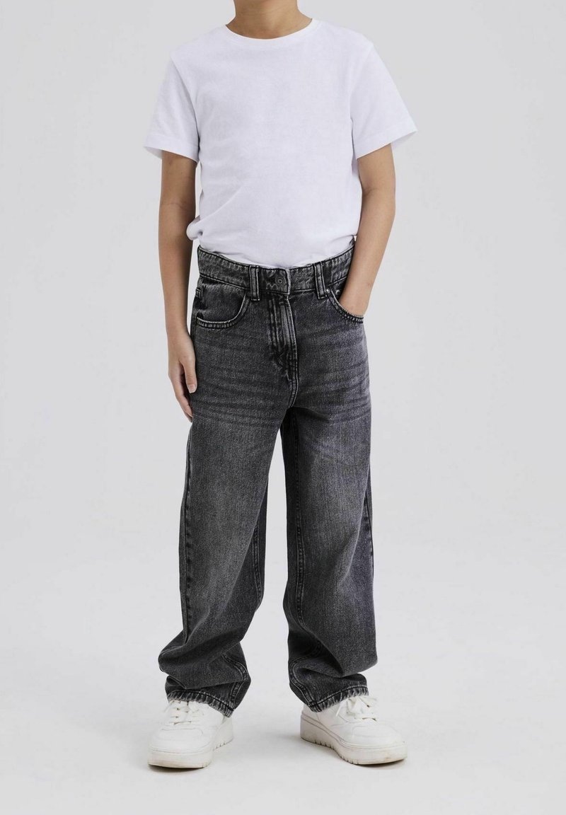 Enfant portant un t-shirt blanc uni, un jean gris foncé ample et des baskets blanches, debout avec une main dans la poche.