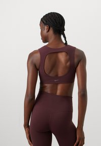 Mørk burgunder sports-bh med en cut-out ryggdesign og Nike-logo. Bh-en har et mykt, elastisk stoff og er kombinert med matchende høyhalsede leggings.
