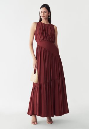 Donna che indossa un abito lungo senza maniche rosso scuro, che tiene una piccola borsa beige, in piedi su uno sfondo bianco uniforme.