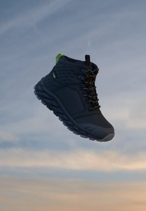 Botte de randonnée noire avec semelle épaisse et languette verte suspendue sur fond de ciel nuageux.