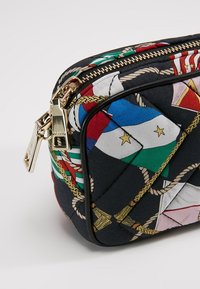 Pochette noire matelassée avec motifs de drapeaux nautiques et tirettes de fermeture éclair dorées sur un fond clair.