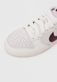 Sapatilha branca de couro com caixa de biqueira perfurada, sola de borracha bege e logótipo da Nike em marrom contrastante. Apresenta atacadores brancos planos e textura suave.