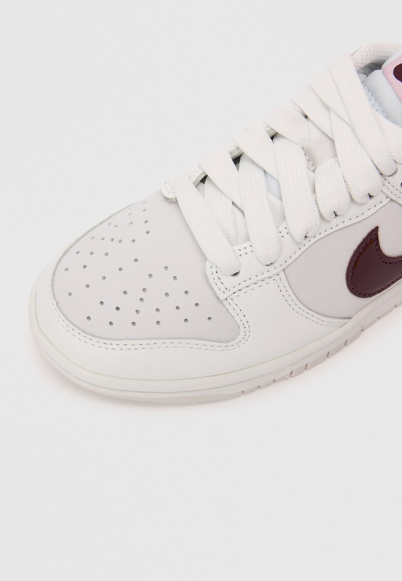 Witte leren sneaker met geperforeerde neus, beige rubberen zool en contrasterend kastanjebruin Nike-logo. Voorzien van platte witte veters en een gladde textuur.