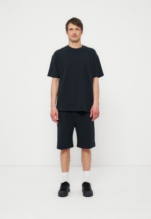 Νέος άνδρας που στέκεται σε λευκό τοίχο, φορώντας μαύρο oversized t-shirt, σορτς μήκους έως το γόνατο στο ίδιο χρώμα, λευκές κάλτσες και μαύρα αθλητικά παπούτσια.